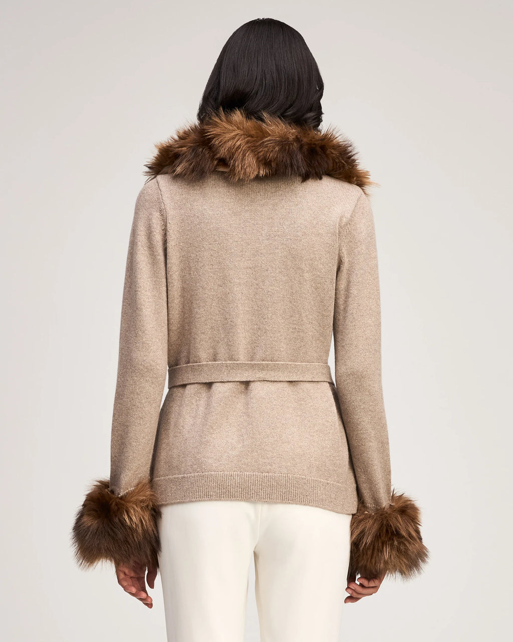 Back view of a model wearing the Cinq à Sept Sandy Cardigan in Oatmeal, showing the full knit drape, self-tie belt back, and fur-trimmed collar.