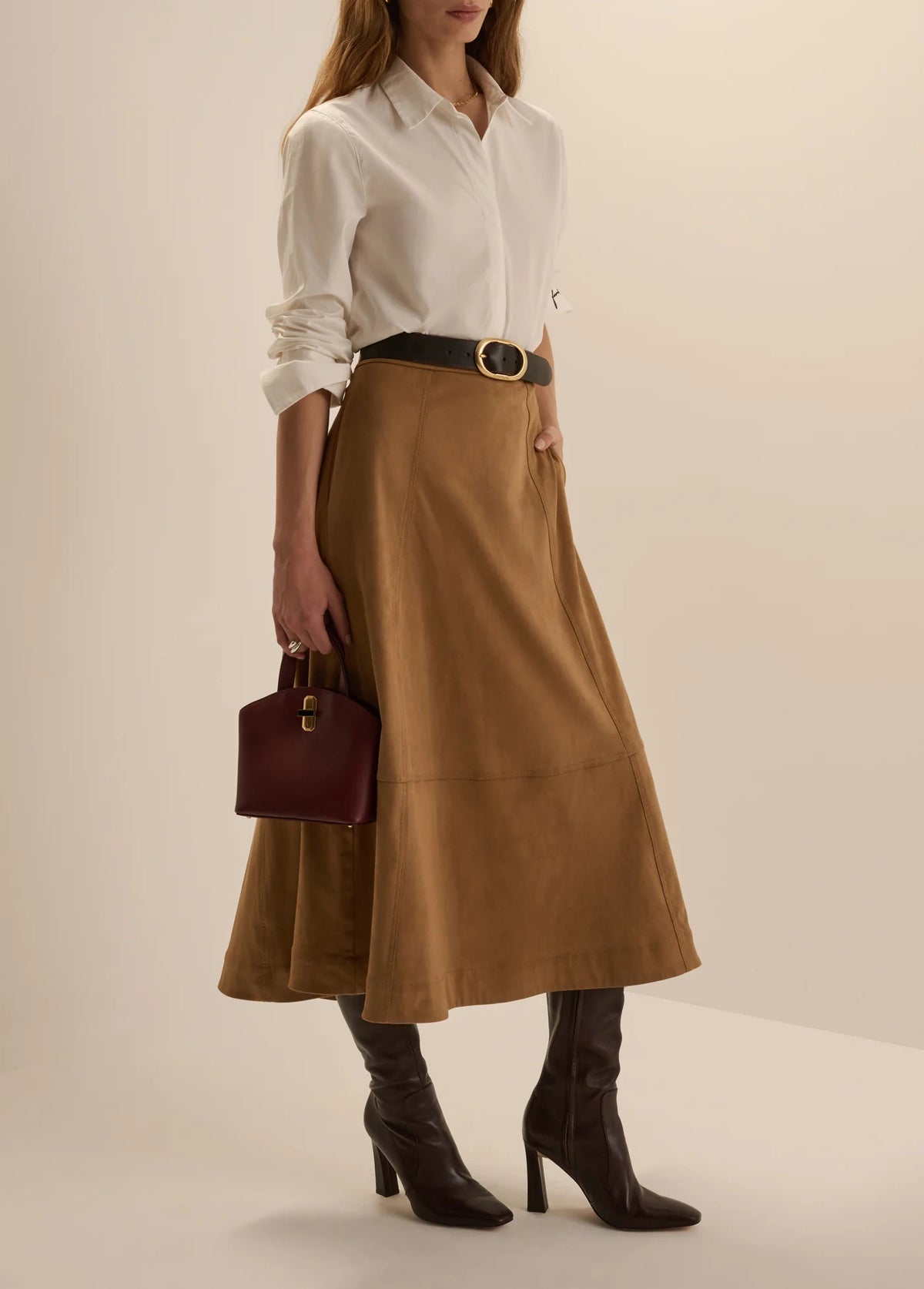 Three-quarter side view of a woman wearing the Favorite Daughter The Aspen Skirt in camel faux suede, styled with a white button-down shirt, a black belt, dark brown high-heeled boots, and carrying a burgundy handbag.