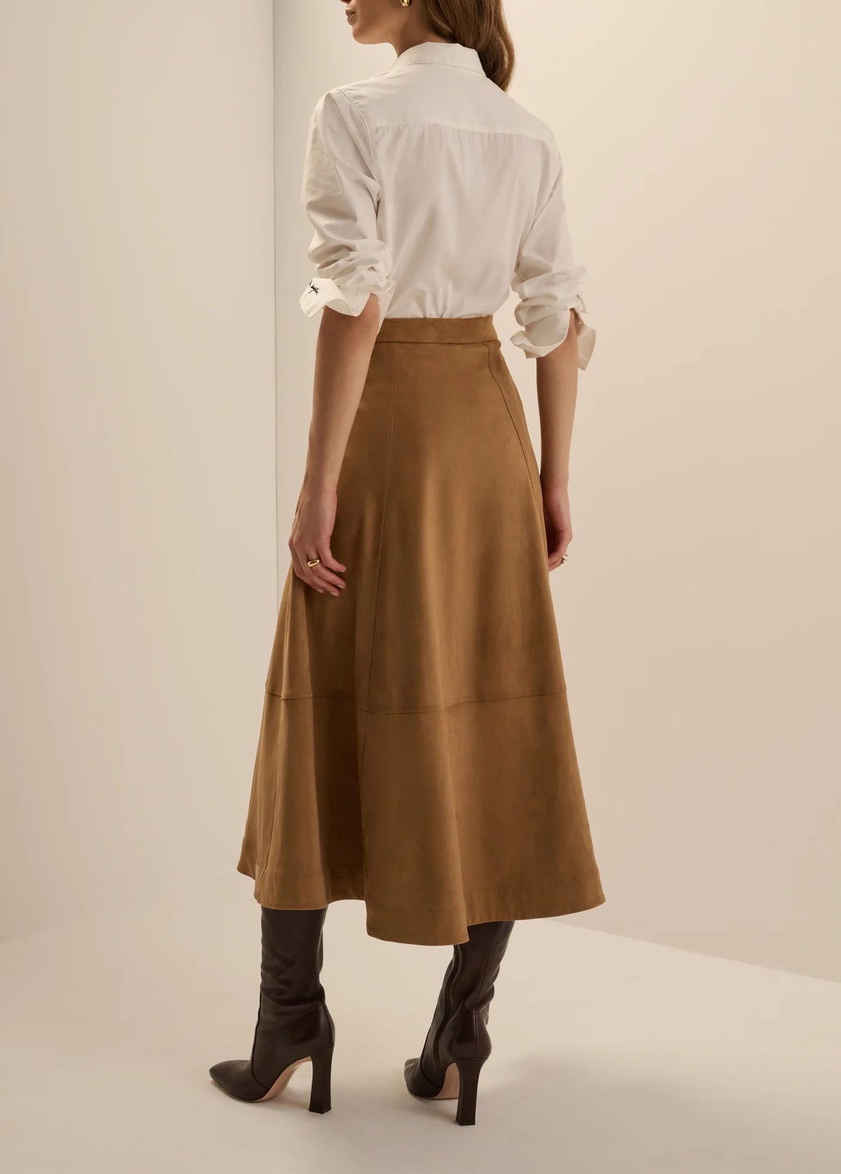 Back view of a woman wearing the Favorite Daughter The Aspen Skirt in camel faux suede, styled with a white button-down shirt and dark brown high-heeled boots.