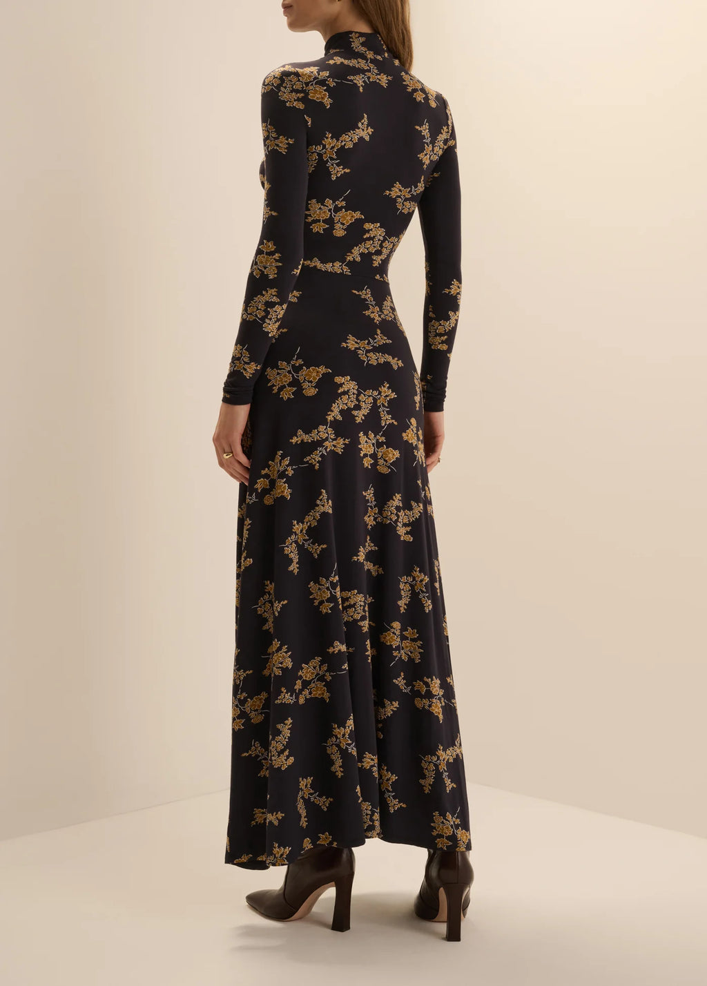 Back view of a woman wearing the Favorite Daughter Lucia high-neck, long-sleeve maxi dress in navy vine floral print, paired with dark brown high-heeled ankle boots.