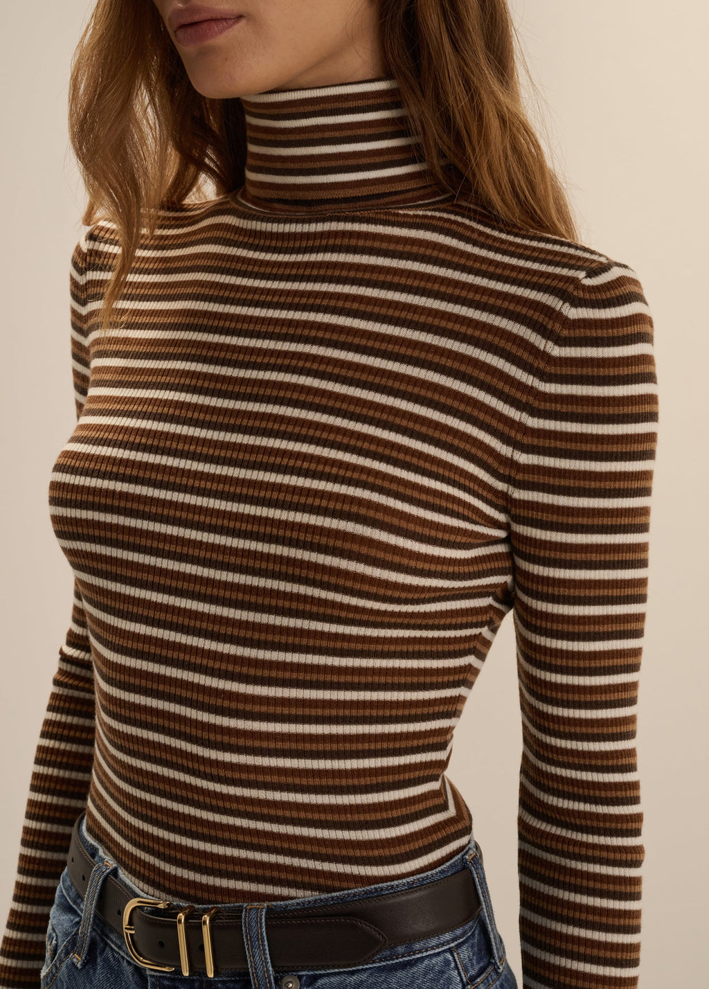 Close-up front view of a woman wearing the Favorite Daughter Amelia Long Sleeve turtleneck top in toffee mini stripe, styled tucked into blue jeans with a black belt.