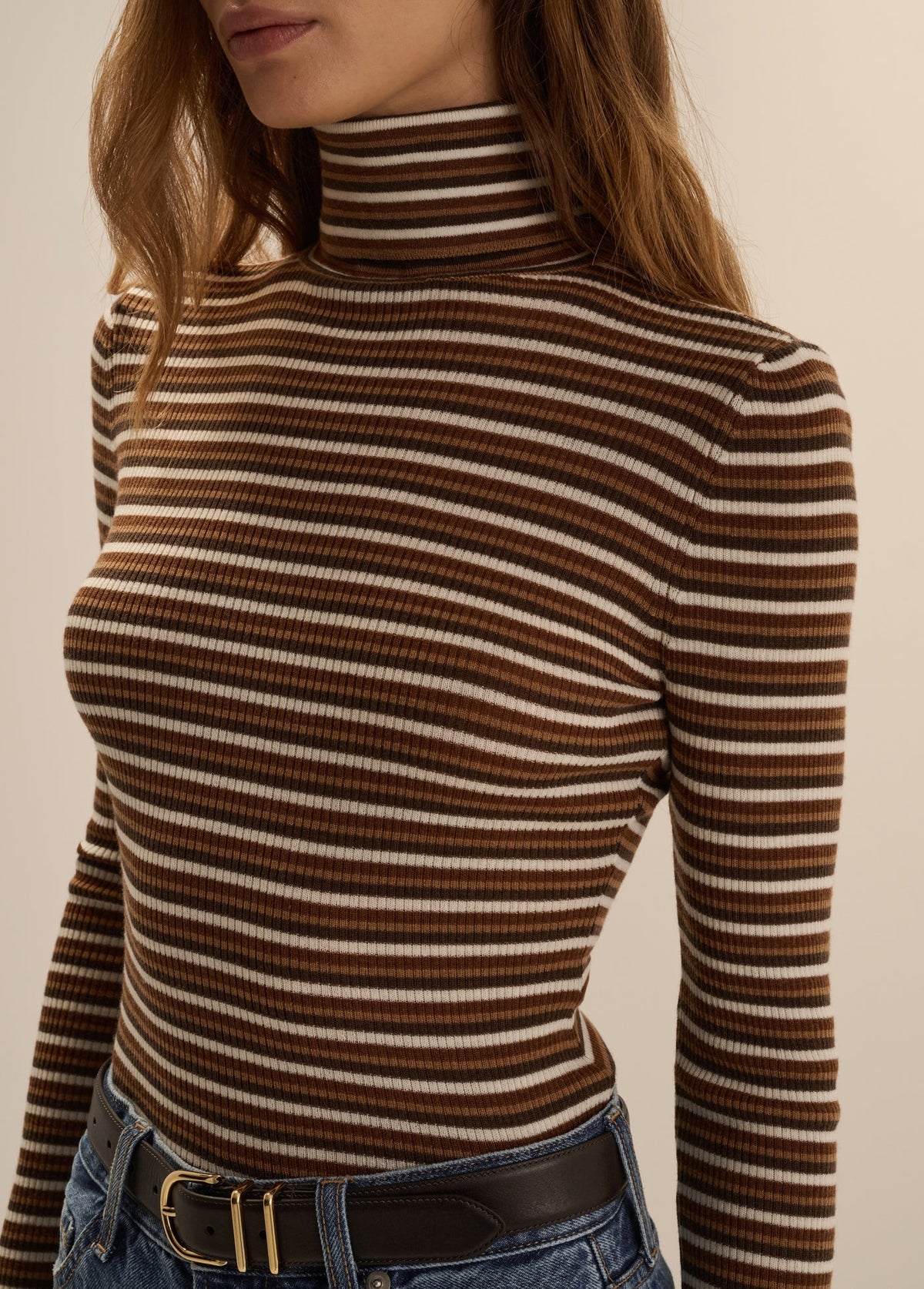 Close-up front view of a woman wearing the Favorite Daughter Amelia Long Sleeve turtleneck top in toffee mini stripe, styled tucked into blue jeans with a black belt.