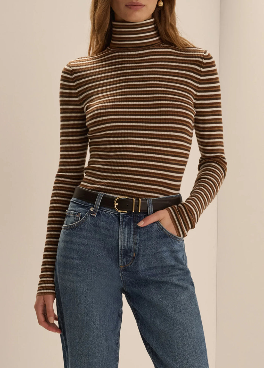 A woman wearing the Favorite Daughter Amelia Long Sleeve turtleneck top in toffee mini stripe, styled tucked into blue jeans with a black belt.