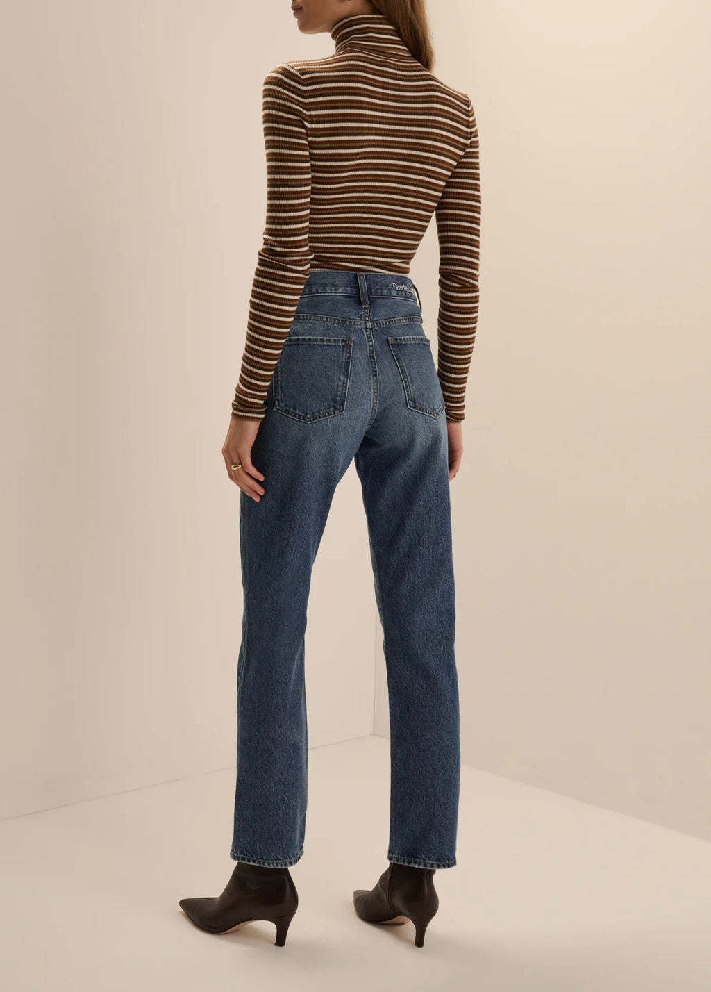 Back view of a woman wearing the Favorite Daughter Amelia Long Sleeve turtleneck top in toffee mini stripe, styled tucked into blue jeans with dark brown kitten-heel ankle boots.